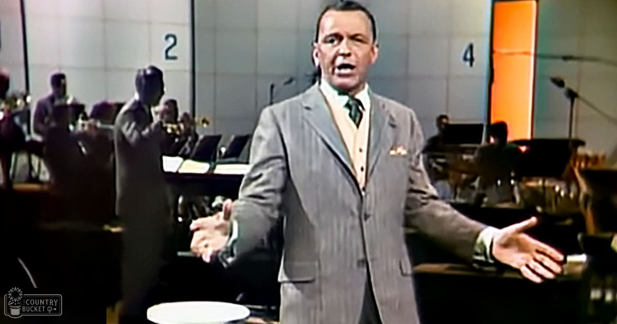 A middle-aged man in a gray pinstripe suit stands with arms outstretched on an indoor stage or studio. Multiple young adults and middle-aged men play trumpets behind him, illuminated by artificial light.