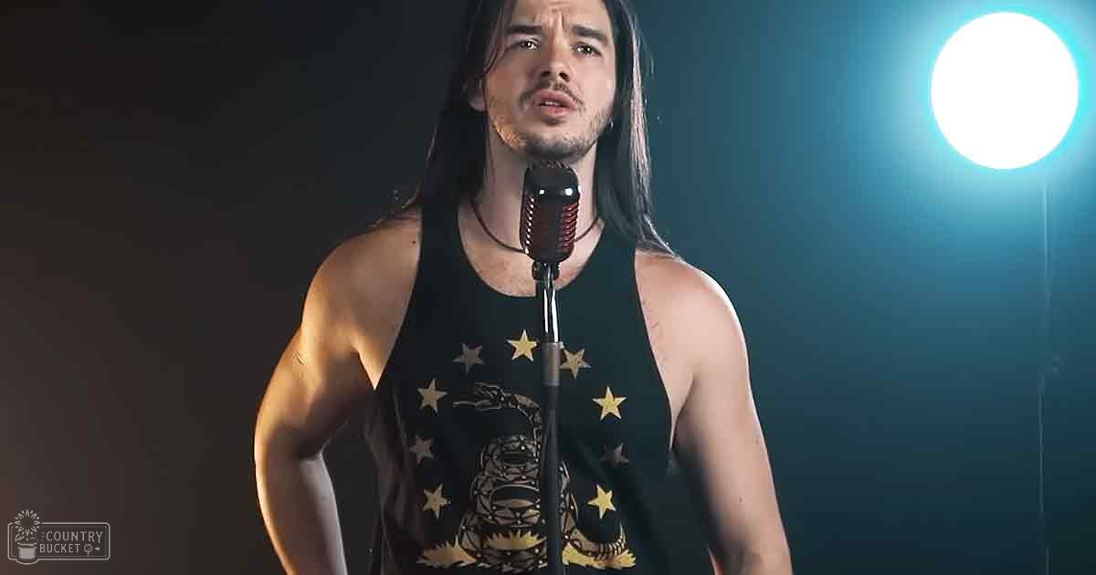 A young adult male singer in a black tank top with gold star and snake design, performing at an indoor music venue under dramatic stage lighting.