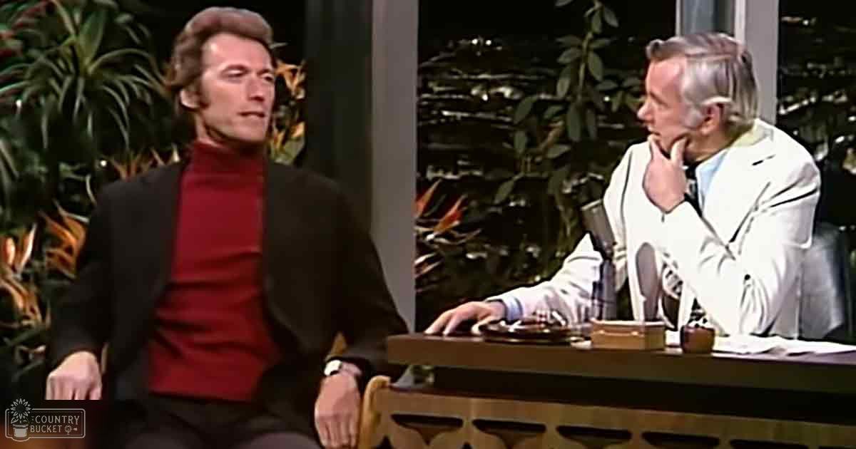 Adult male guest in dark blazer over red turtleneck on stage with older adult male wearing white suit jacket against backdrop of night cityscape, urban setting at spring or summer evening