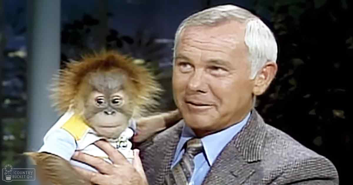 An older adult male wearing a patterned blazer and light blue shirt holds a juvenile Bornean orangutan in a studio setting, likely for an educational or talk show program.