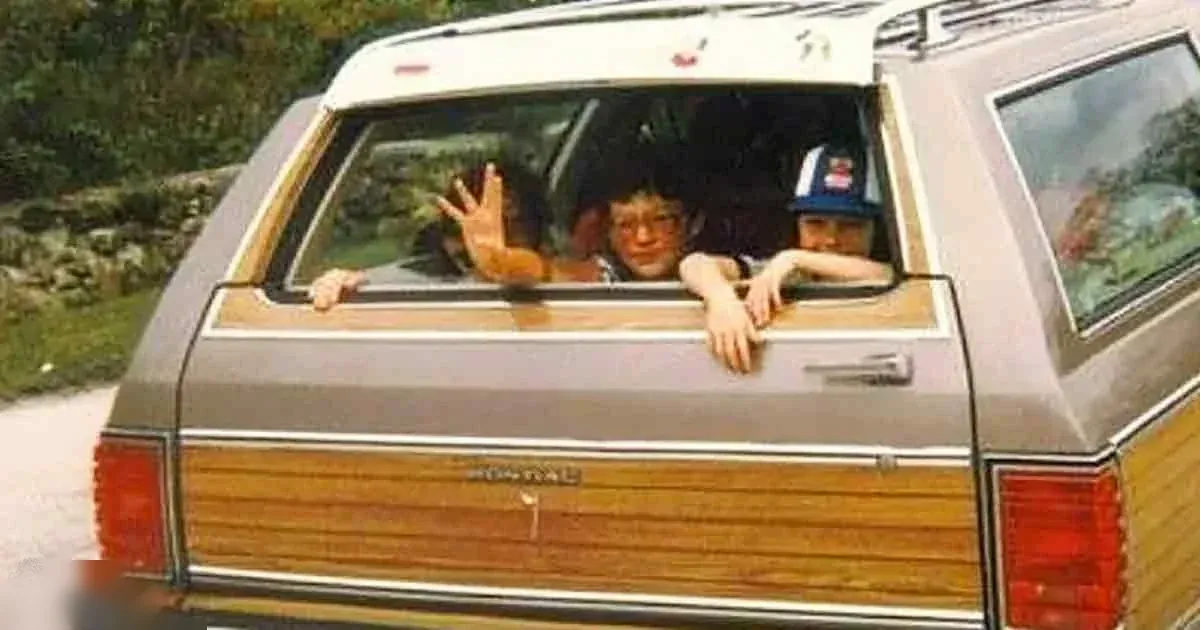 A family of two children in a station wagon with wooden paneling, outdoors amidst greenery during daytime.