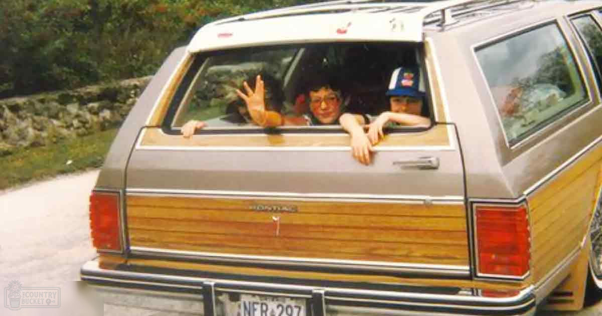A vintage station wagon with two children leaning out of the rear window in a rural area during midday.