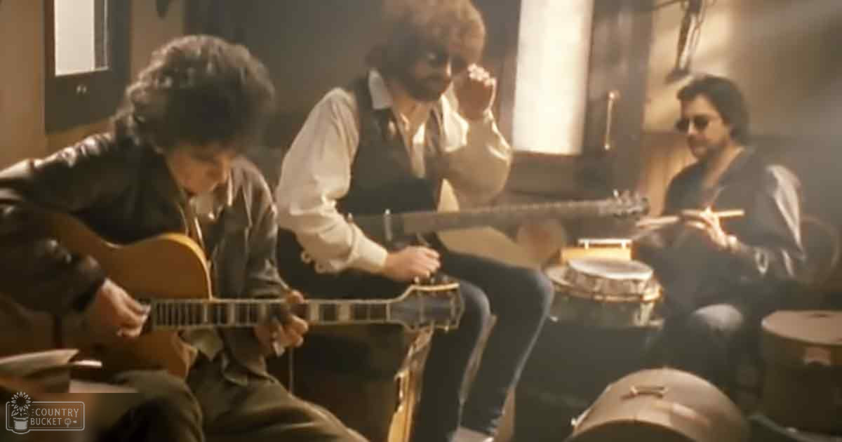 Musical performance scene indoors, young adult males wearing casual clothing with a drum set and stringed instruments in natural light.