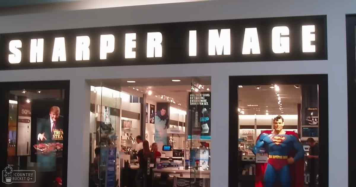 A middle-aged adult male wearing a white shirt is inside a store front labeled "SHARPER IMAGE" with various items displayed on shelves and a Superman action figure in the window. The setting appears to be indoors, likely within a shopping mall.