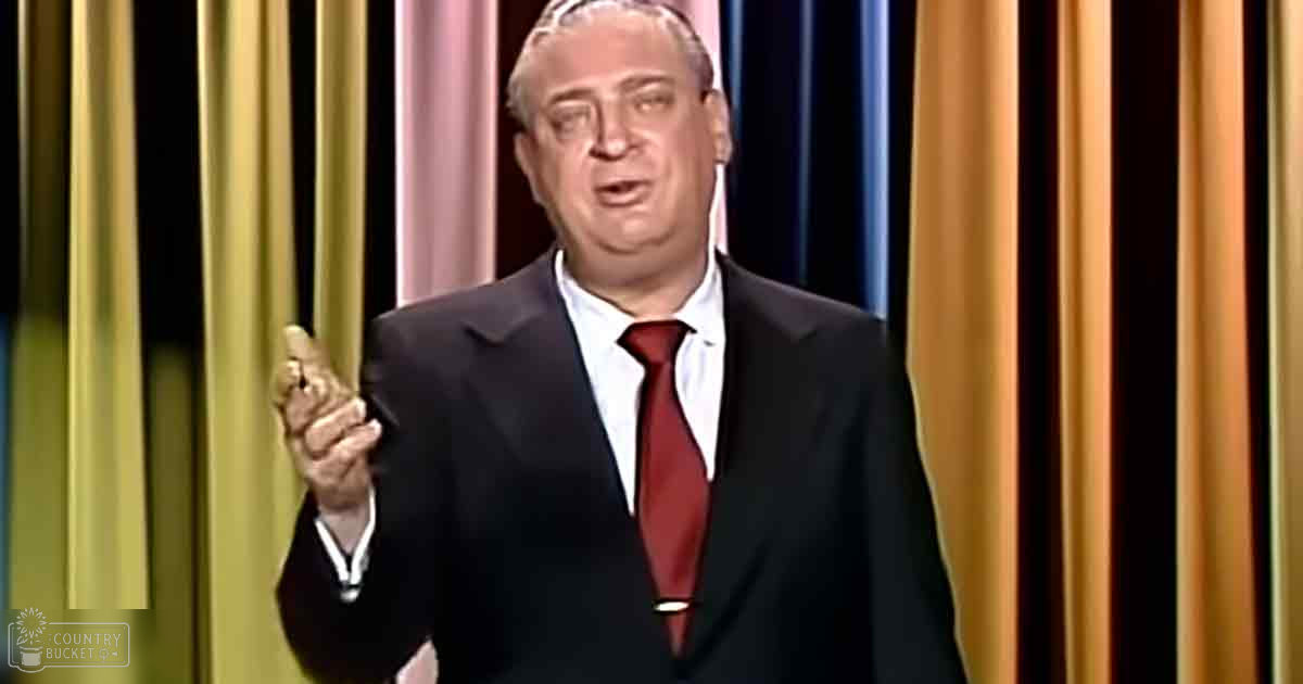 A middle-aged adult male in a formal suit with black jacket, white collared shirt, and red tie stands against vertical striped curtains of gold, pink, blue, and orange on an indoor stage-like setting.