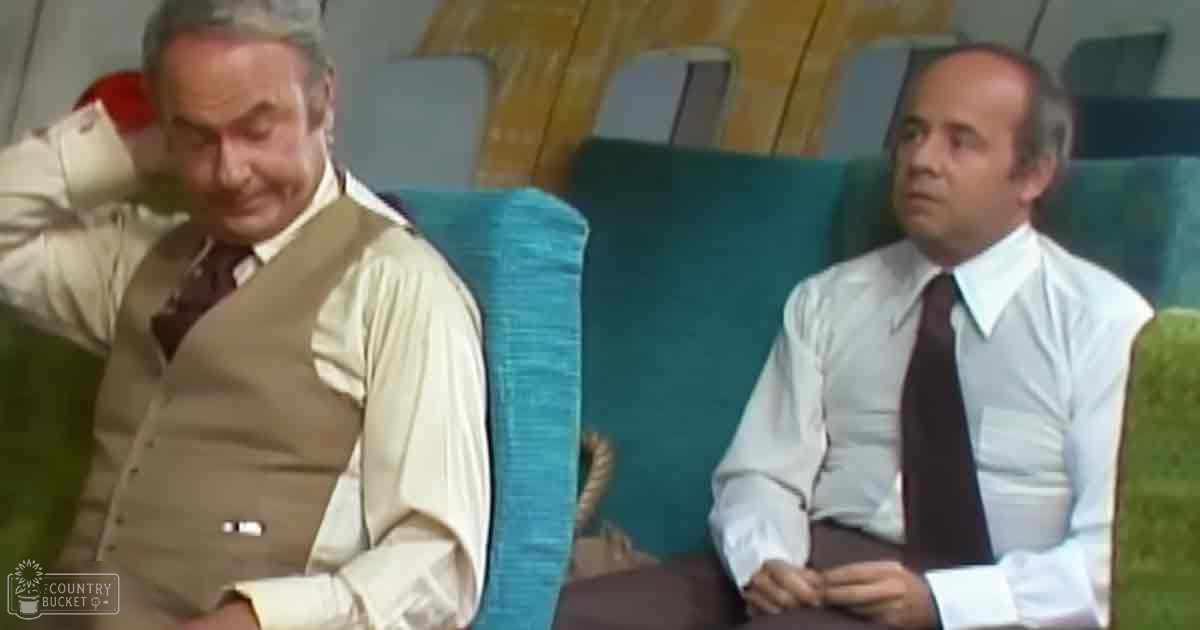 Two middle-aged men seated in an indoor room, possibly an office or living space; teal chairs with wooden armrests and a wooden wall behind them.