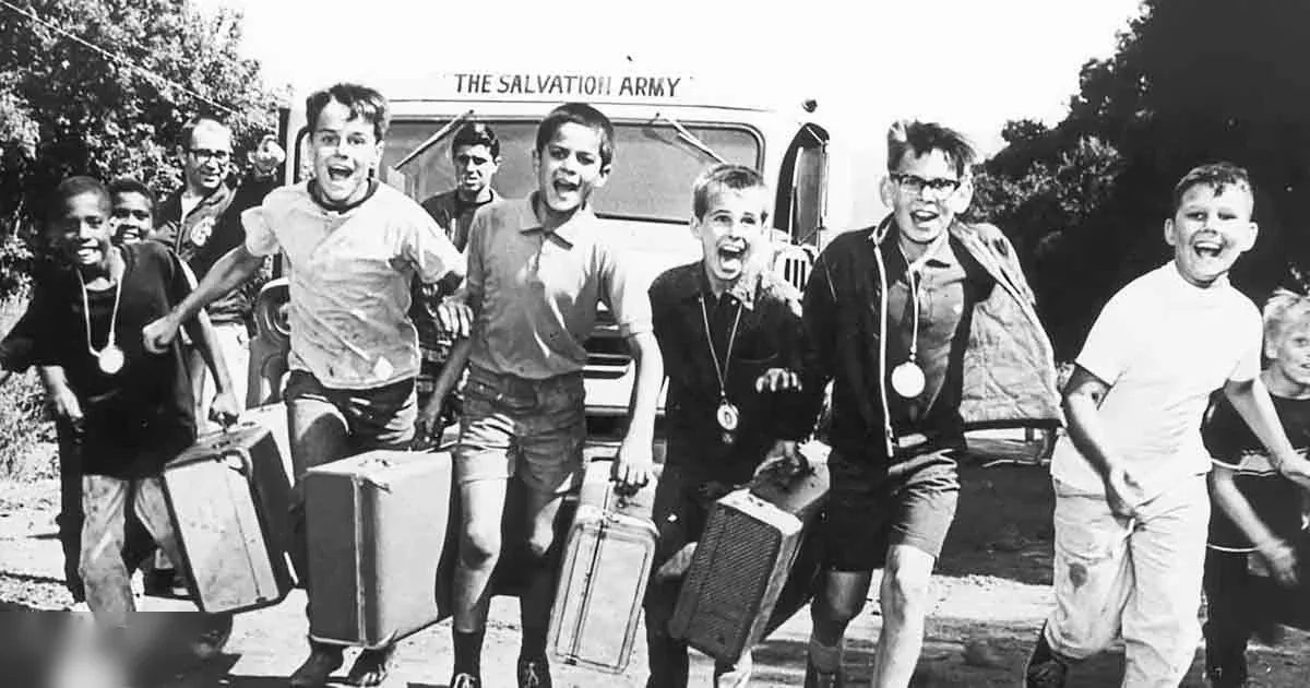 A group of children (ages 8-14) stand outdoors near a Salvation Army vehicle with trees in the background, suggesting late afternoon light.