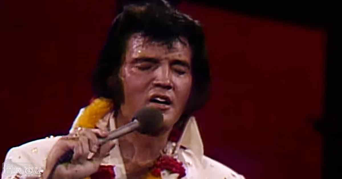 An adult male singer in a form-fitting, white shirt with gold embellishments and red accents, performing on stage under artificial lighting.