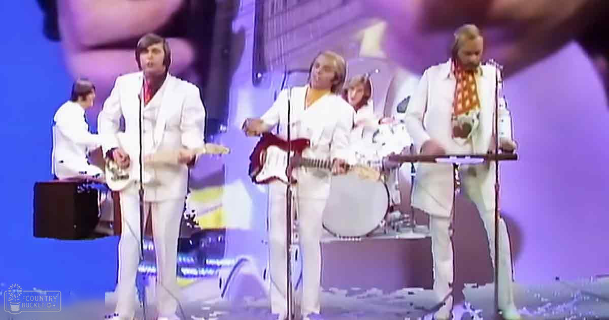 A musical performance on a stage with performers in white suits, red shirts, and a keyboard player wearing an orange patterned scarf. The setting is indoors under artificial lighting.