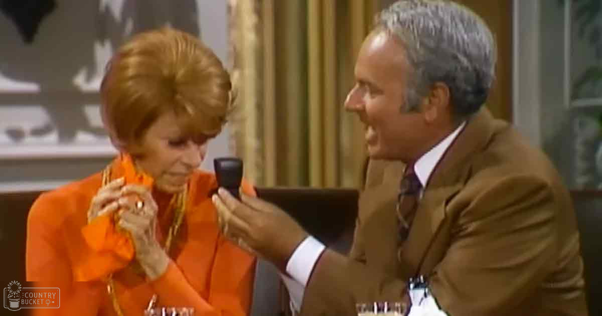 Two older adults in a talk show setting, one wearing a bright orange vintage-style outfit with ruffled details and a patterned scarf, the other in a brown suit jacket over a white shirt with a dark tie, holding a microphone against a backdrop of curtains and stage elements.