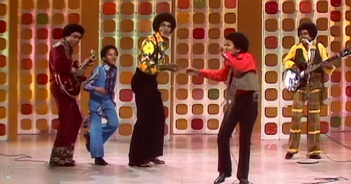 Adult male singer in maroon suit, young adult in blue checkered jumpsuit, and other performers on stage with retro aesthetic and colorful wall background.