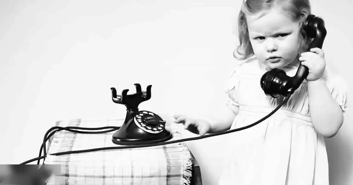 A toddler wearing a simple white dress indoors, accompanied by a vintage rotary telephone on a plain background.