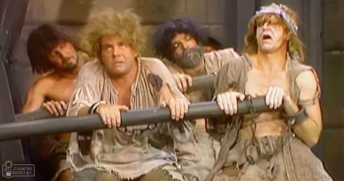 A theatrical scene with three adult males in loose, tattered attire indoors under bright lighting.