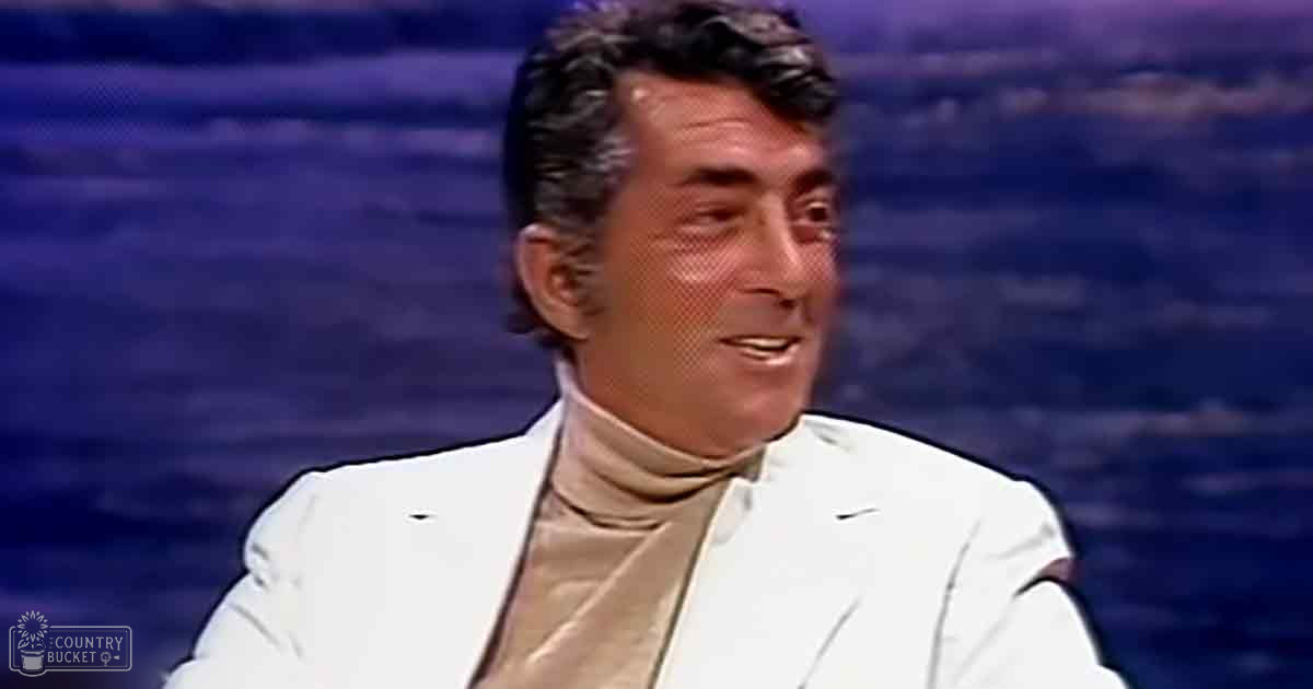 An adult male wearing a white blazer and beige turtleneck in a studio setting with dark background, likely from a television production.
