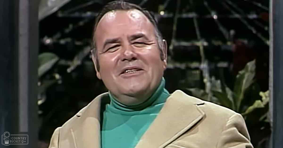 An older adult male in a studio setting, wearing a beige trench coat with teal turtleneck underneath.