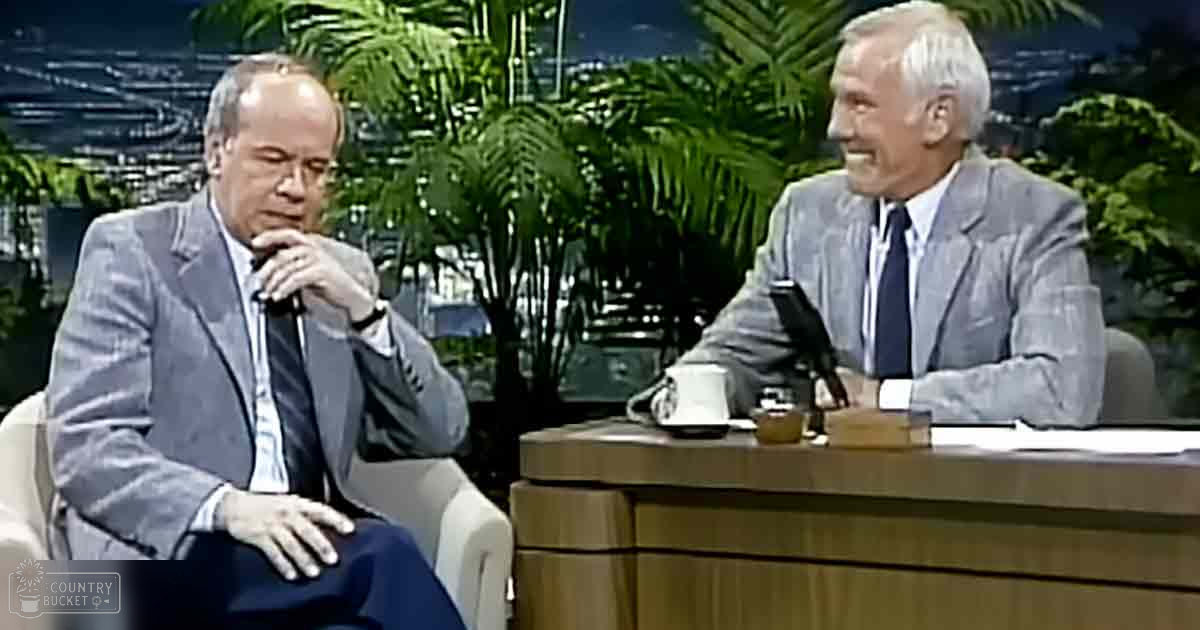 Two older adults engaged in conversation within a talk show setting; muted greens of plants, dark blues and grays of suits, whites and light grays of shirts, indoor urban scene with professional lighting.