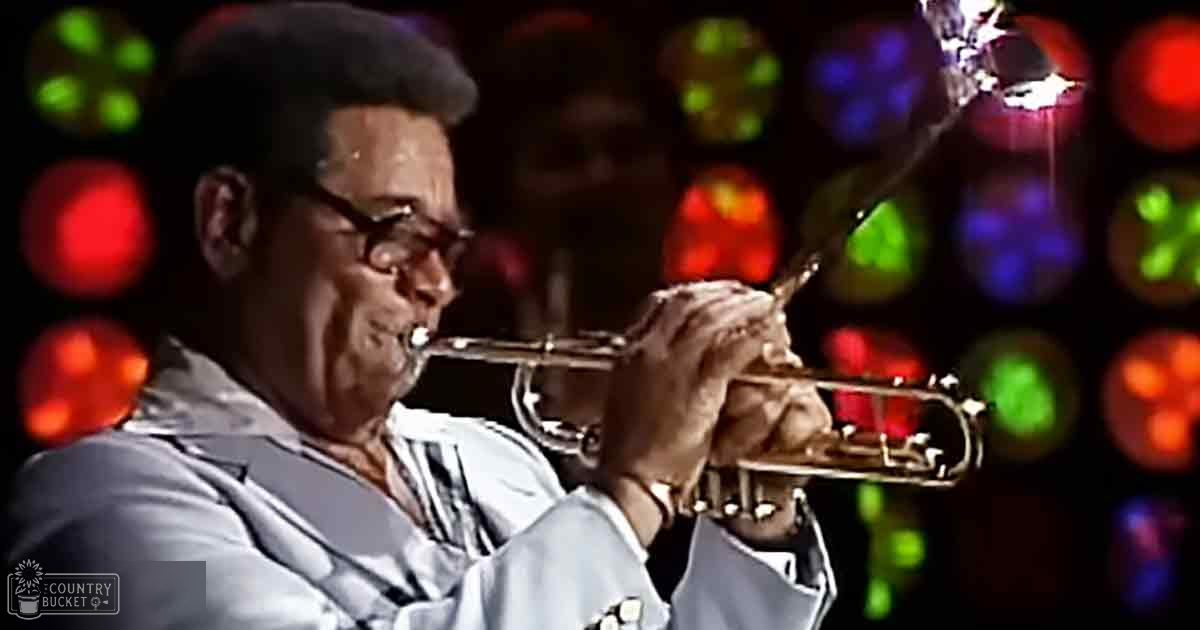 An older adult male trumpet player wearing a light-colored striped suit jacket and dark trousers performs on stage under colorful bokeh lighting.