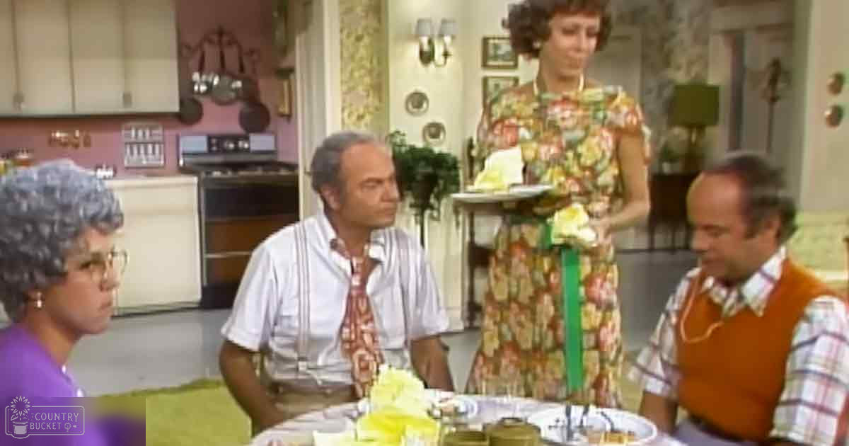A woman in a floral dress holds a tray with food items; an older adult female wearing glasses and a purple garment stands to the left, while two middle-aged males in white shirts and orange vests are present. The scene is set indoors, likely in a home kitchen with mid-20th-century decor.