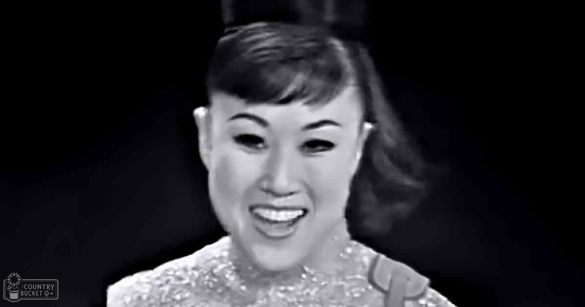 An adult female in a fitted, embroidered dress from the mid-20th century against an indoor backdrop with artificial lighting.