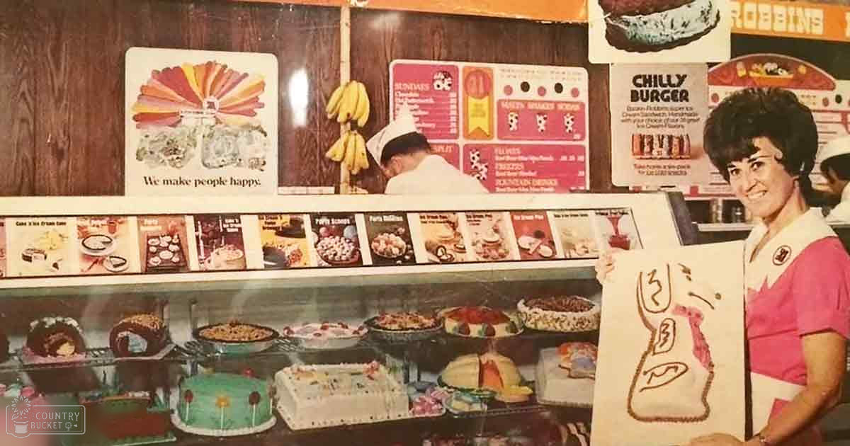 Adult female holds paper with drawing; adult male in chef's hat behind counter. Bakery display case filled with cakes, pastries, and menu board listing 'Chilly Burger' and other food options.