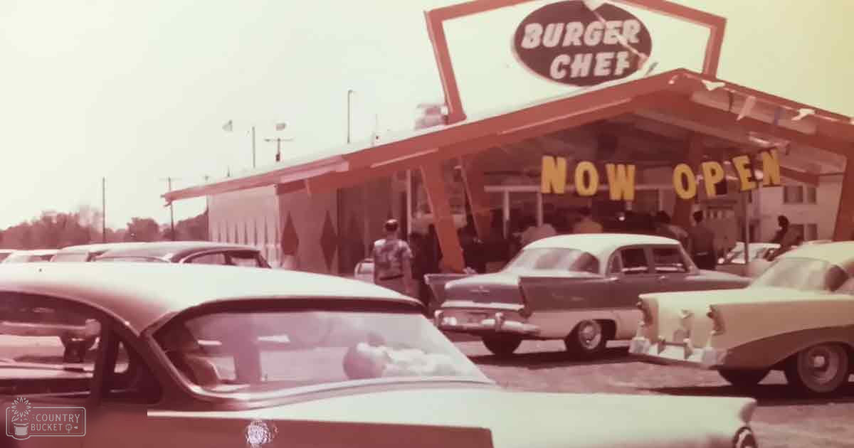 An adult male singer in a light-colored shirt stands near Burger Chef restaurant entrance with another person, vintage cars parked outside under clear skies.