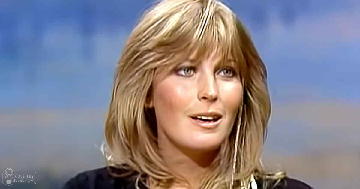 A middle-aged female in an indoor setting, wearing a dark, simple cut garment from the 1970s or early 1980s with large metallic earrings.