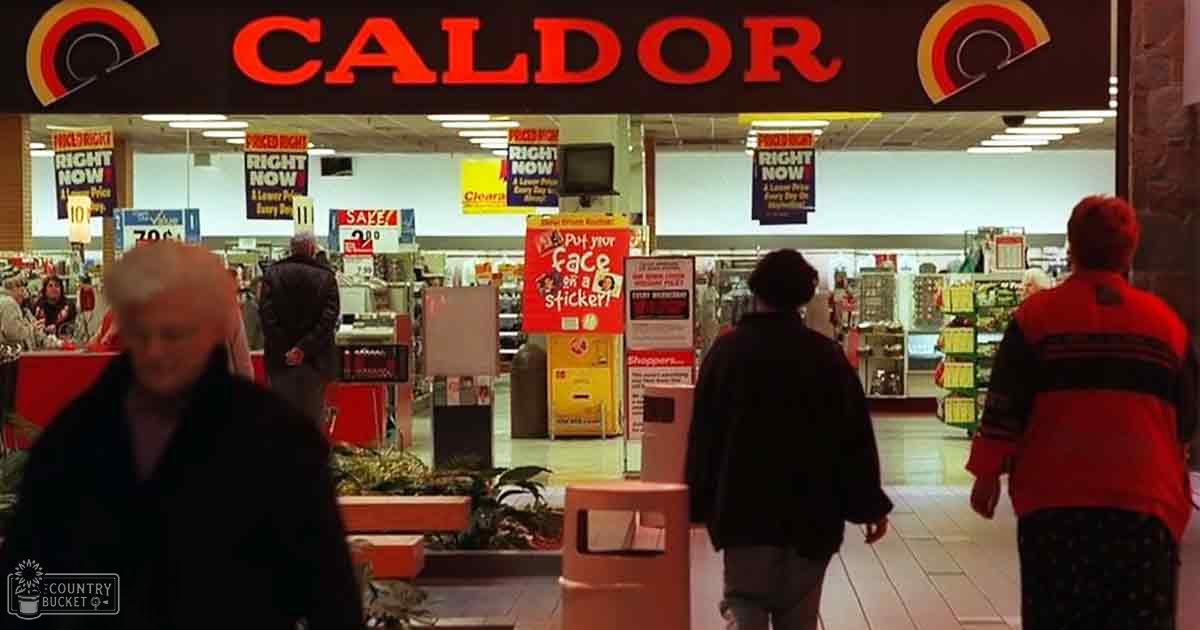 Caldor store interior from 1980s-early 2000s with older female in dark coat and adult male or female in dark jacket, red and black striped sweater, various promotional banners and signage visible.