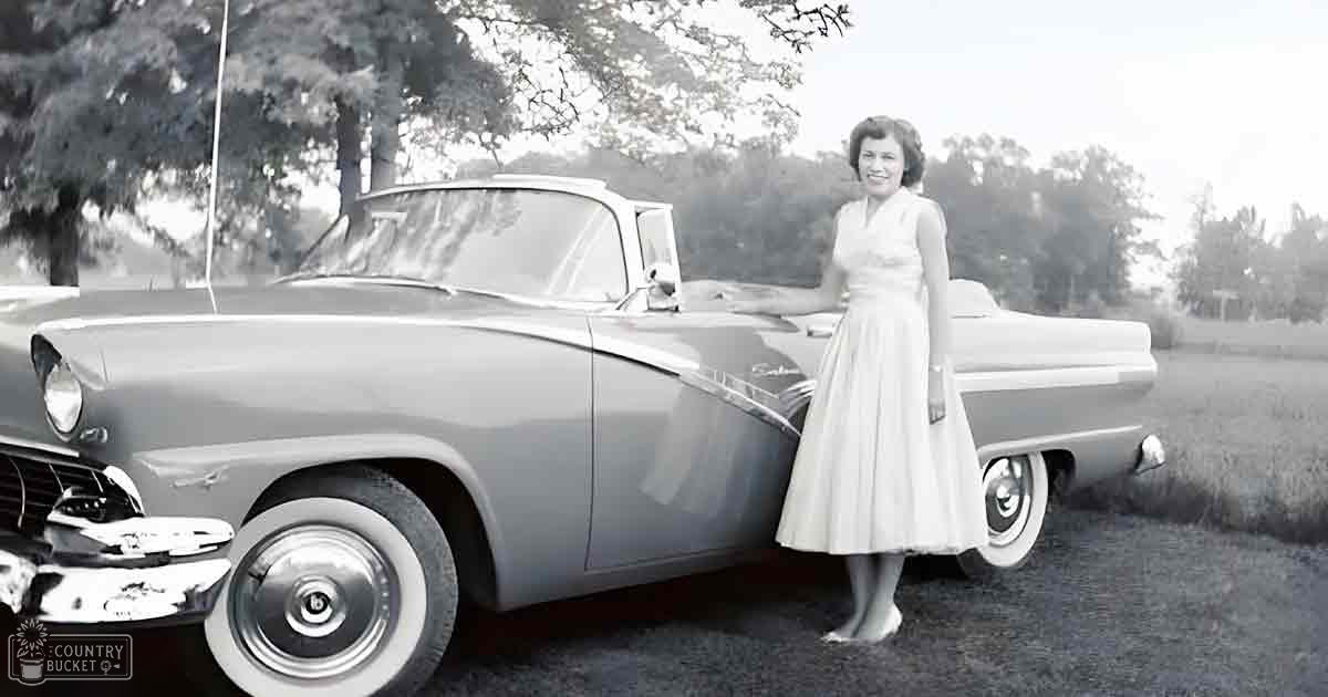 A woman in a white mid-20th-century dress stands next to a classic car with chrome detailing, set against a backdrop of trees and grassy fields under natural daylight.
