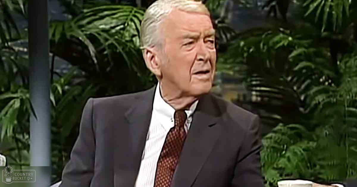 An older adult male wearing a formal dark gray suit and white collared shirt with red tie, standing against an indoor garden backdrop.