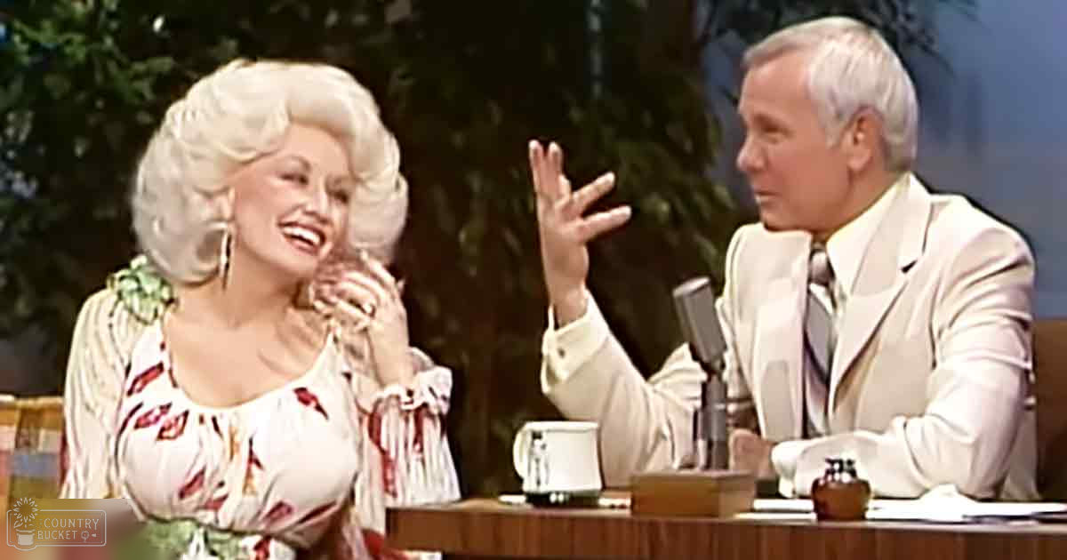 A woman wearing a floral dress and bouffant wig sits next to an older male host on a talk show set.