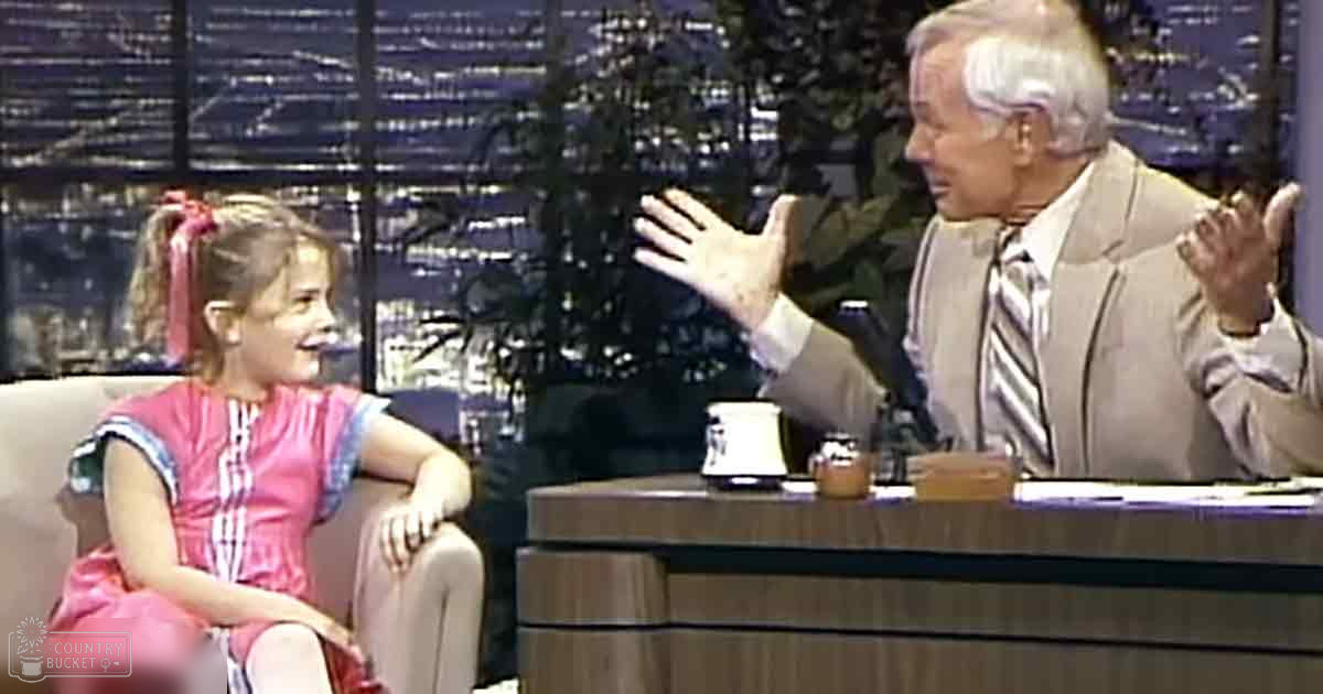 A child in a pink striped dress with red hair tied up, seated on an armchair next to an adult male behind a desk at a talk show set with a cityscape backdrop.
