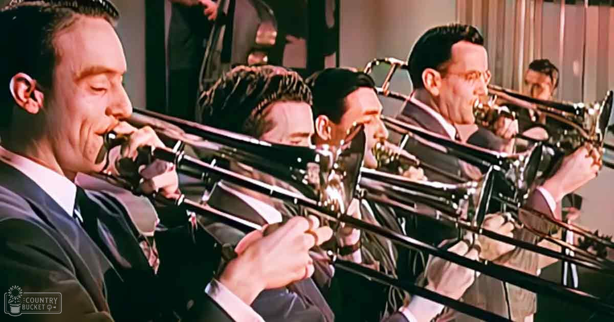 A group of adult male musicians playing trombones on a stage-like setting with dark suits and white shirts, curtains or drapes in the background suggesting an indoor venue.