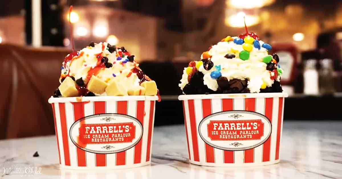 Two ice cream sundaes with whipped cream, chocolate sauce, M&Ms, and other toppings on red and white striped cups at Farrell's Ice Cream Parlour Restaurants in a warm-lit restaurant setting.