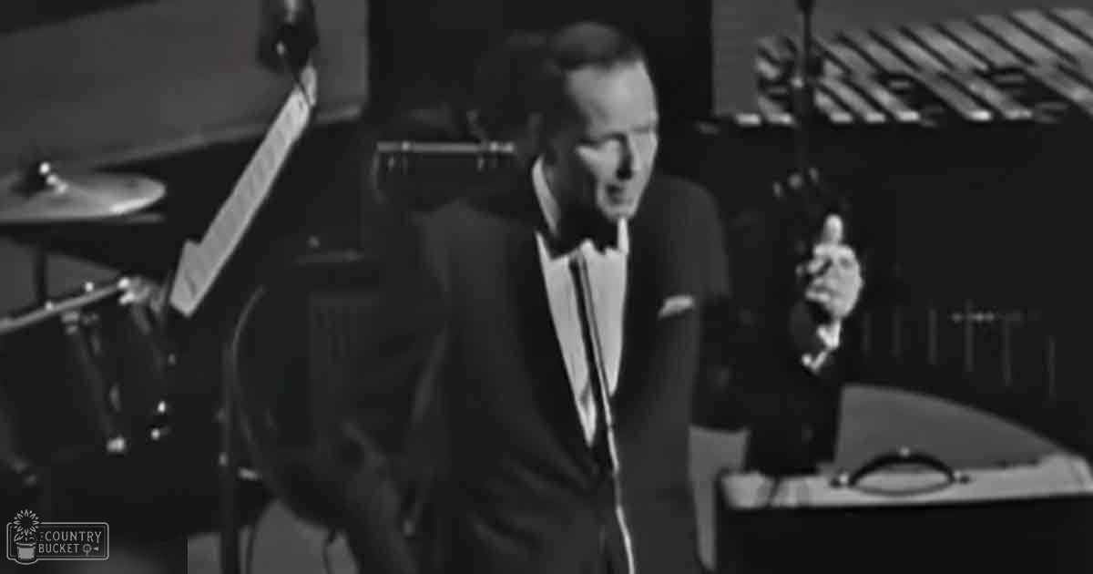 A middle-aged man in a dark suit with a white shirt and tie performs on stage at what appears to be a concert or live music event.
