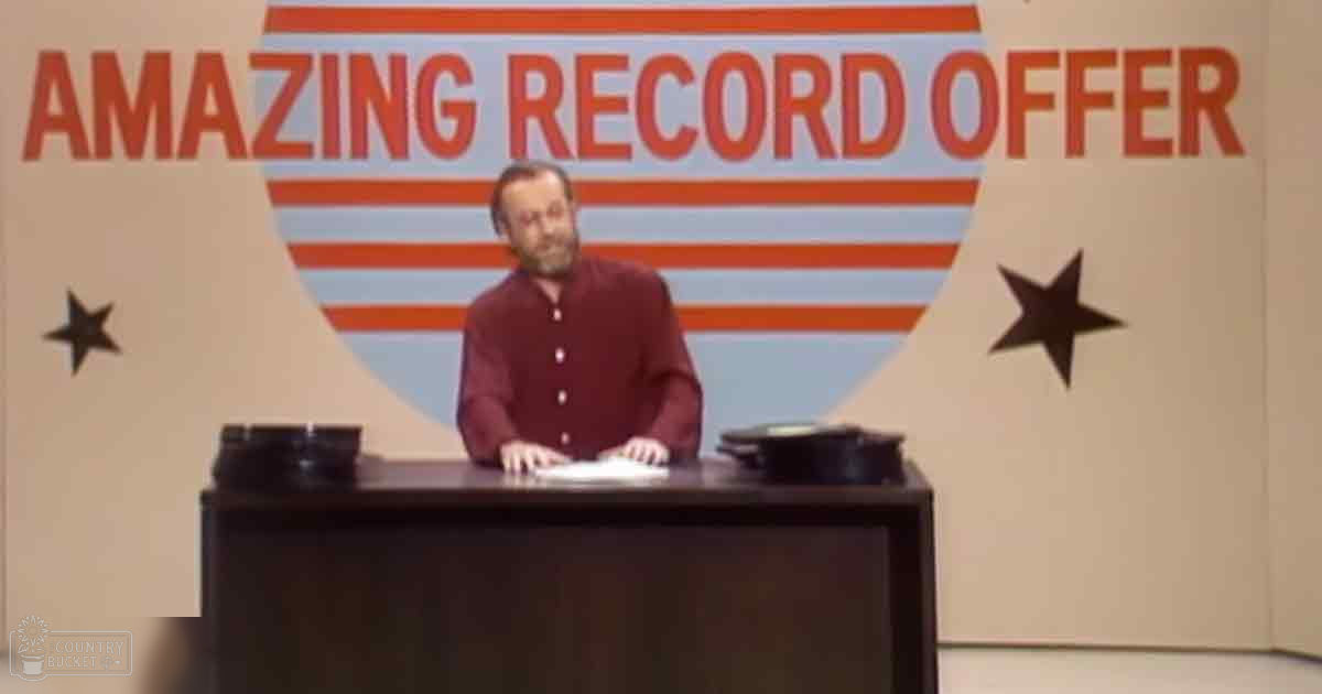 A middle-aged man in a maroon button-up shirt stands before a backdrop advertising 'AMAZING RECORD OFFER,' with two record players on a dark wooden desk.