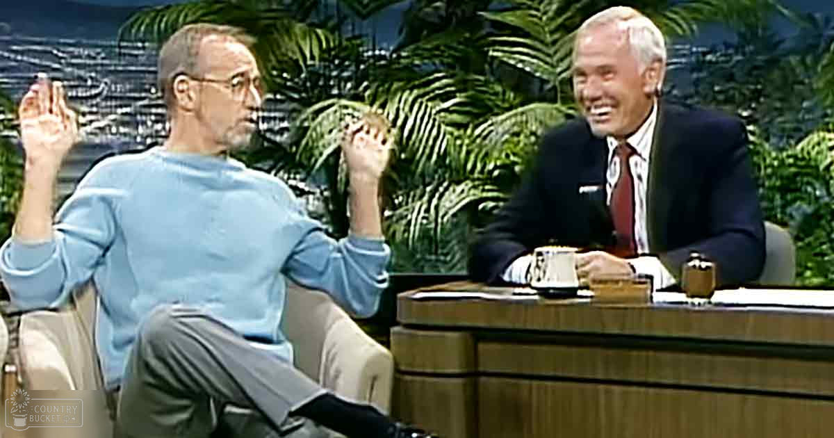 A talk show host dressed in a light blue long-sleeve shirt and gray pants, interacting with an older adult male in a dark suit jacket and white dress shirt on a talk show set.