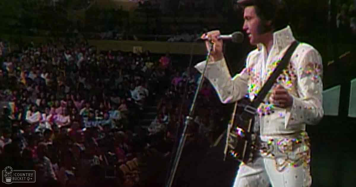 A male adult singer wearing a white sequined jumpsuit performs on stage at an indoor arena during what appears to be a concert.