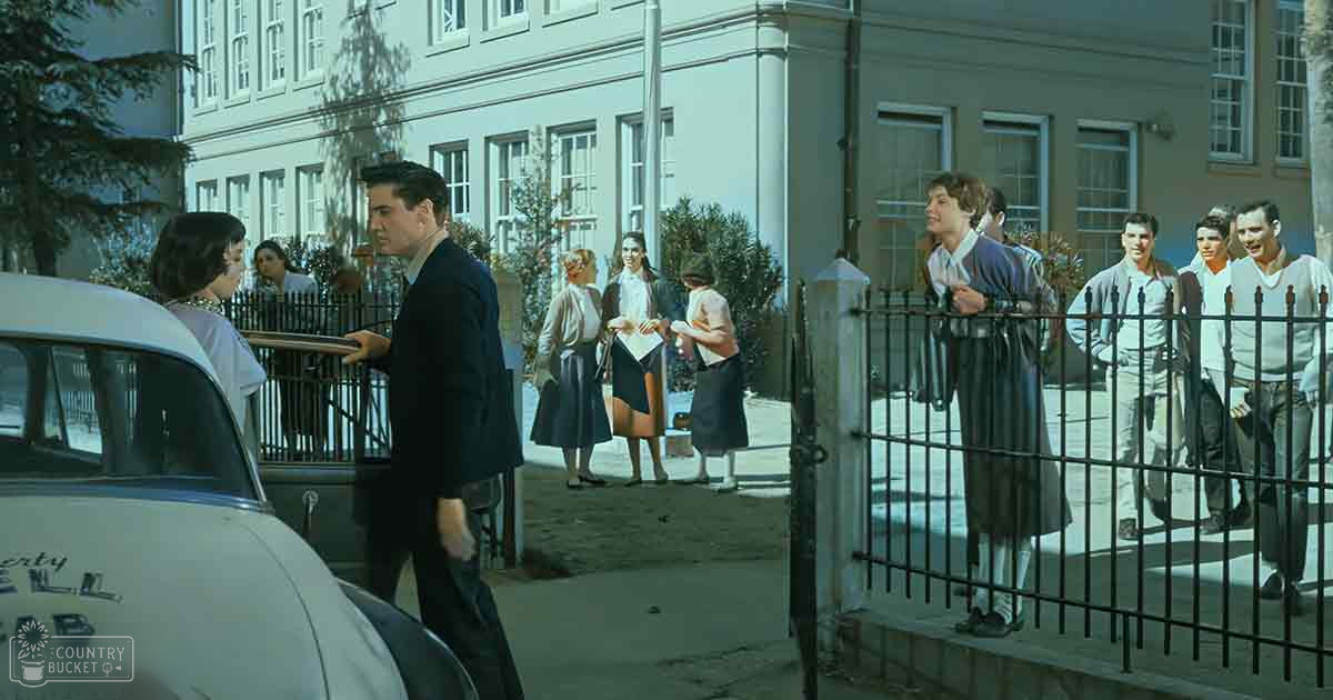 Adult male singer interacting with another individual near a vintage Liberty Bell Cab outside an institutional building, possibly a school or college, on what seems to be a sunny spring day.