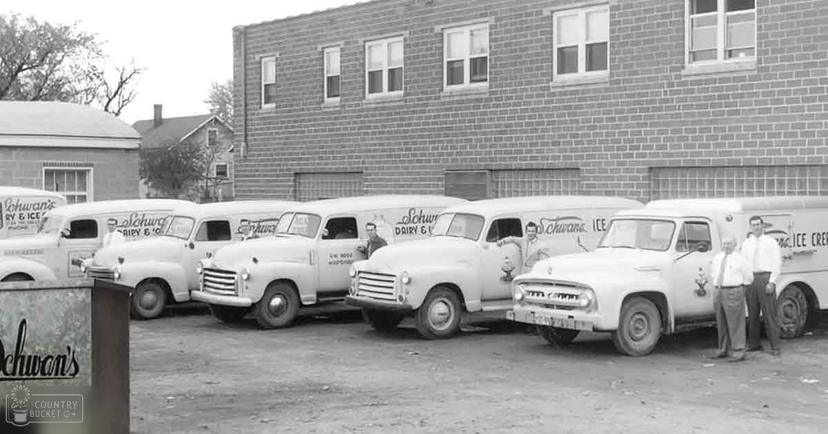 Two men stand beside vintage delivery trucks from Schwan's Dairy & Ice Cream, set against a brick building backdrop in an industrial area with bare trees suggesting autumn.