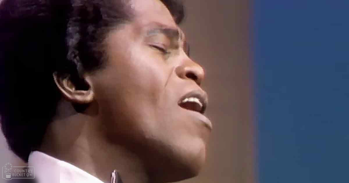 Young adult male singing indoors in a well-fitted white collared shirt with dark tie, mid-20th century style; blurred backdrop suggesting indoor performance venue.