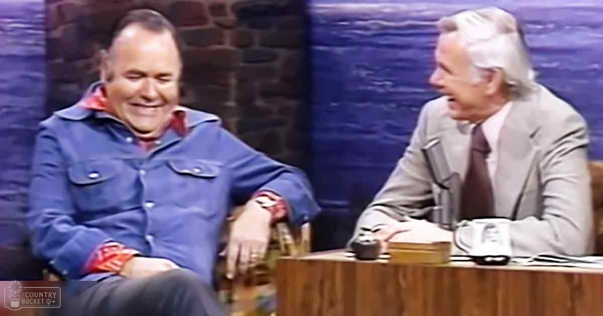 Talk show setting with two individuals in conversation; mid-20th century attire of middle-aged man in blue button-up shirt, older adult man in light gray suit jacket over white shirt and dark tie; studio setup with brick walls and blue curtains, typical of television talk shows from the mid-20th century