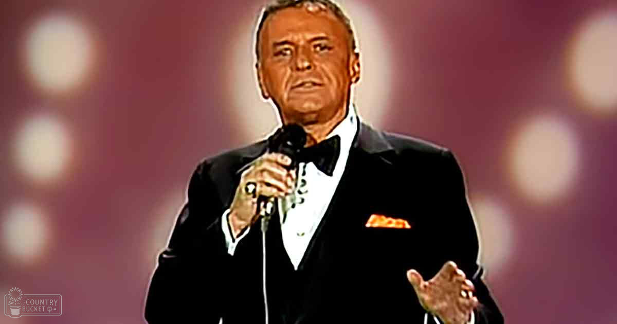A middle-aged adult male wearing a classic black tuxedo with a white shirt and bow tie stands on stage under bright lights at an evening event.