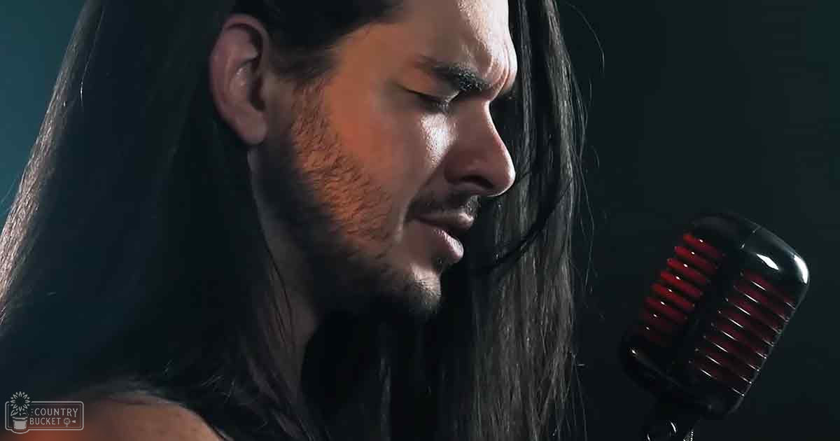 An adult male singer wearing a dark-colored loose-fitting shirt in an indoor setting, singing into a vintage-style microphone with red accents.