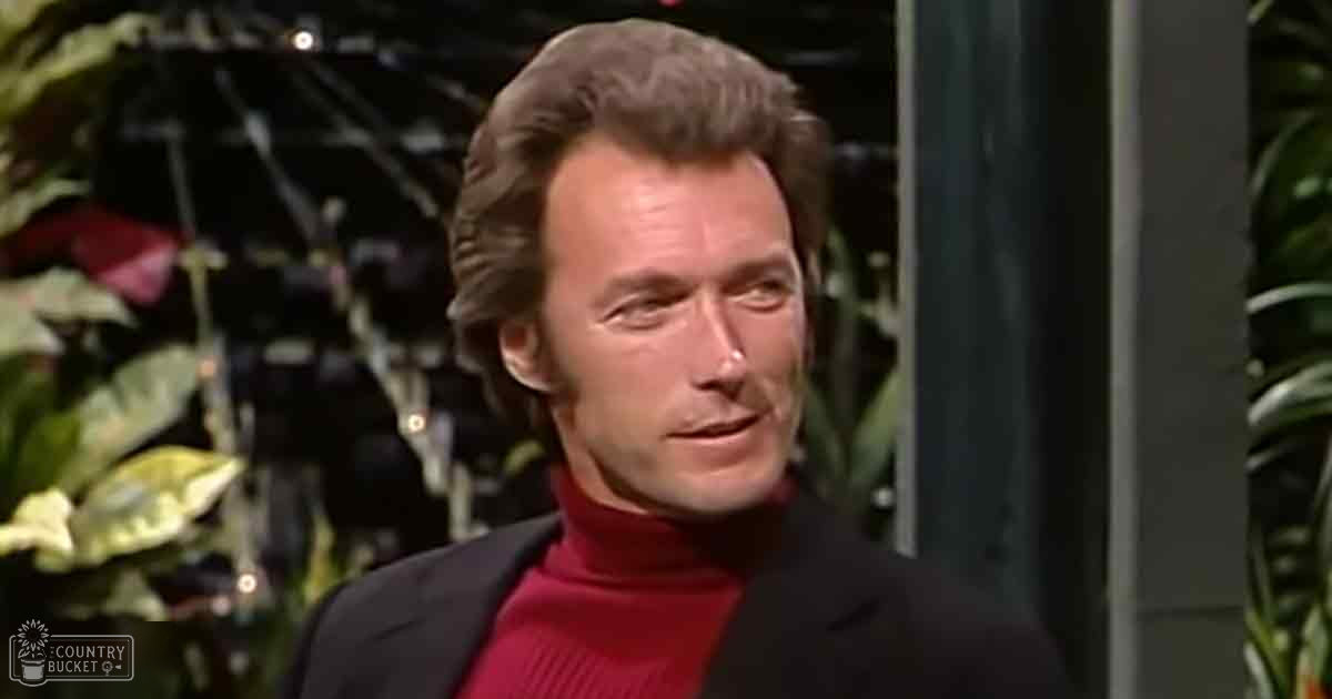 A middle-aged adult male wearing a dark blazer with high collar, red turtleneck sweater, in an indoor setting with decorative plants and stage-like elements.