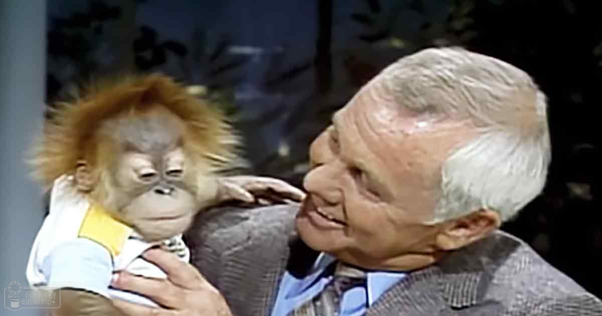 A juvenile orangutan being held in a studio setting with an older adult male wearing a gray suit jacket over a blue collared shirt and patterned tie.