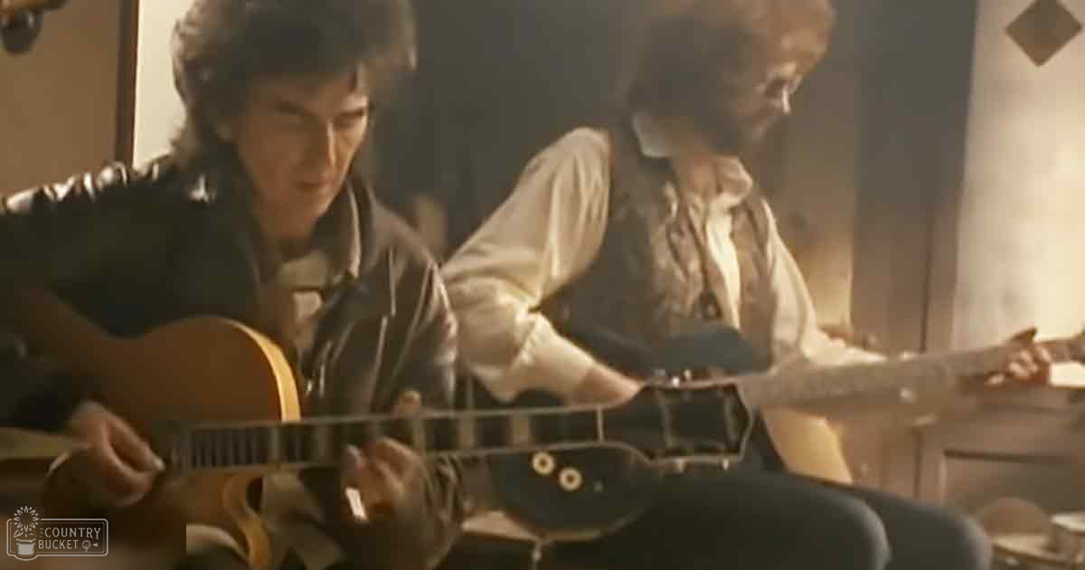 A young adult male playing an acoustic guitar in a dimly lit room with furniture and wall decorations.