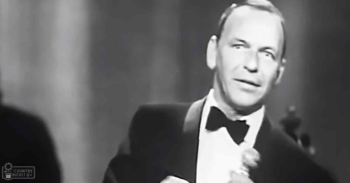 A middle-aged man wearing a formal tuxedo holding a microphone indoors, possibly at an event or performance.