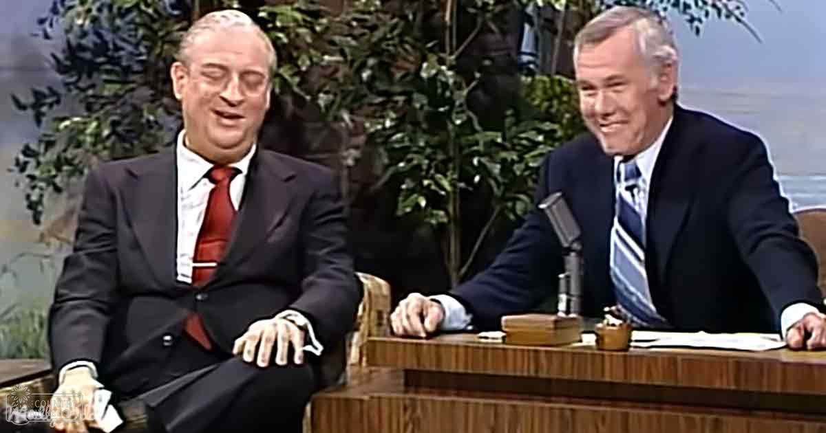 Two men seated at a talk show desk, wearing dark suits and ties with greenery in the background suggesting an indoor studio setting.