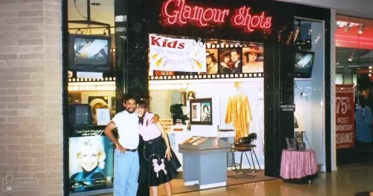 An adult male singer wearing light blue jeans, white t-shirt with dark pants; young female in black skirt with dog design pattern, pink top. Glamour Shots sign visible at mall, table covered with pink cloth.