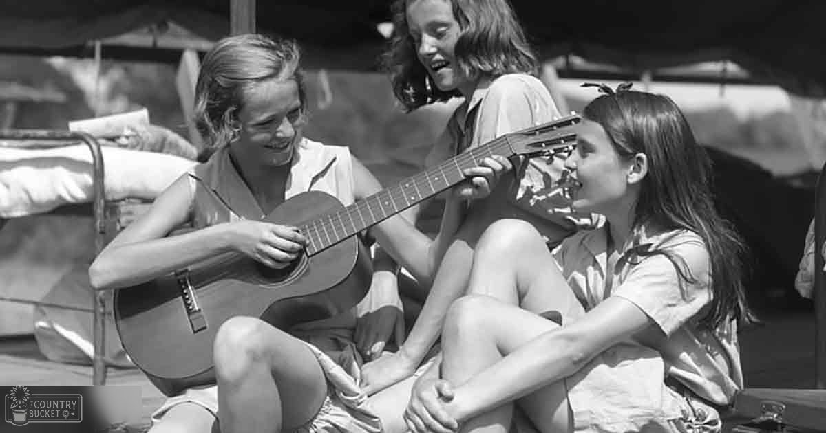 Young adults enjoying music outdoors; a guitar being played by one person with a chair partially visible behind another.